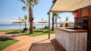 Una terraza de madera con zona de bar, palmeras y sombrillas, con vista a un cuerpo de agua.