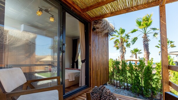 Terraza de madera con puerta de vidrio que da a una habitación con cama y baño. Hay sillas y una pequeña mesa con piñas.