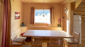 Un interior de madera con una larga mesa de comedor, cuatro sillas, un banco, una ventana con cortinas, una lámpara colgante y una planta pequeña.