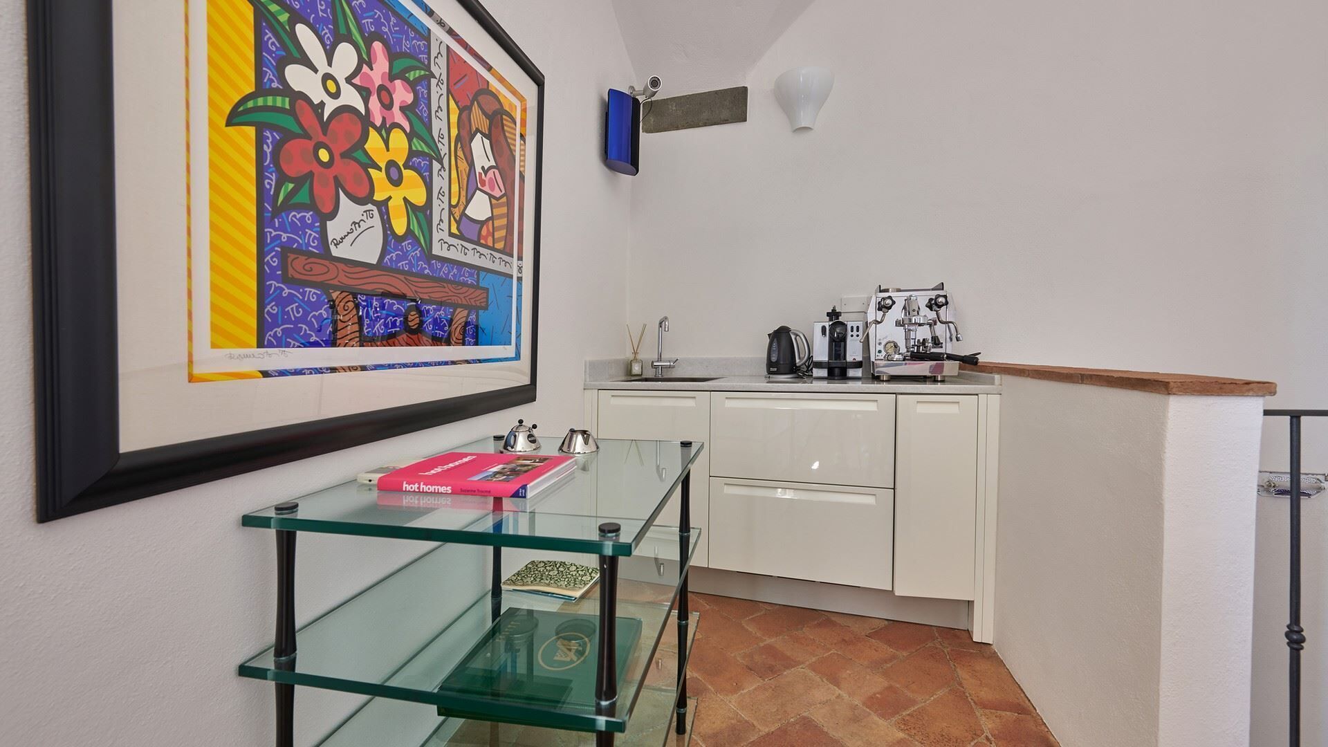A kitchen with a coffee machine, a sink, and a glass shelf with books.