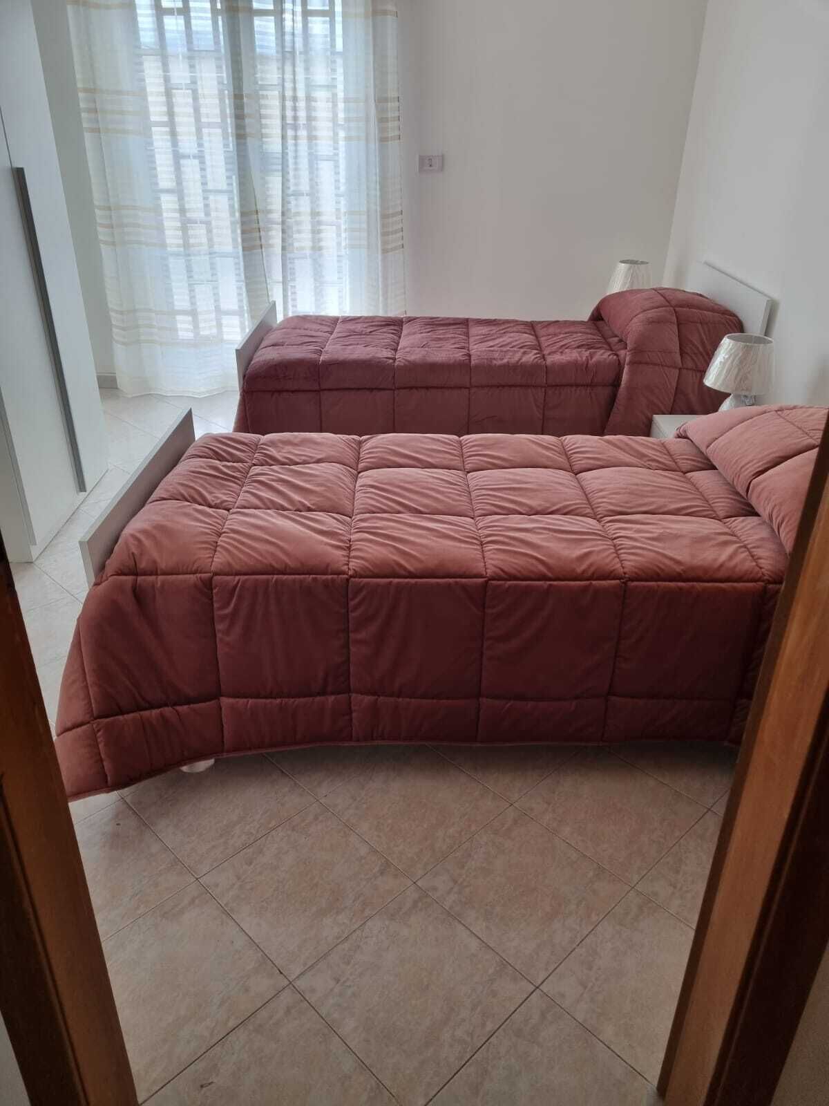 A bedroom with two beds, a window with sheer curtains, and a lamp on a bedside table.