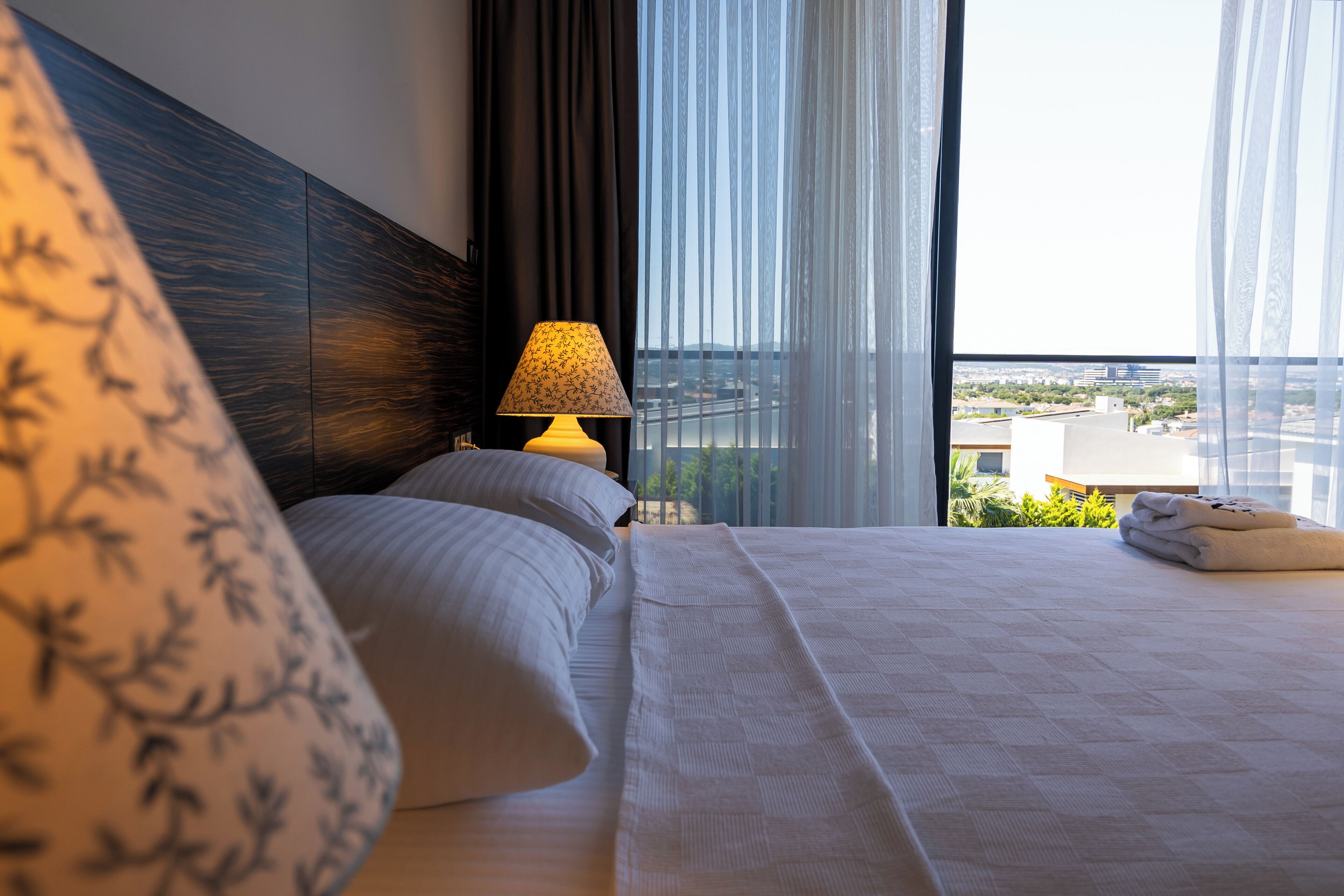 A hotel room with a large bed, a nightstand with a lamp, and a view of the city through sheer curtains.