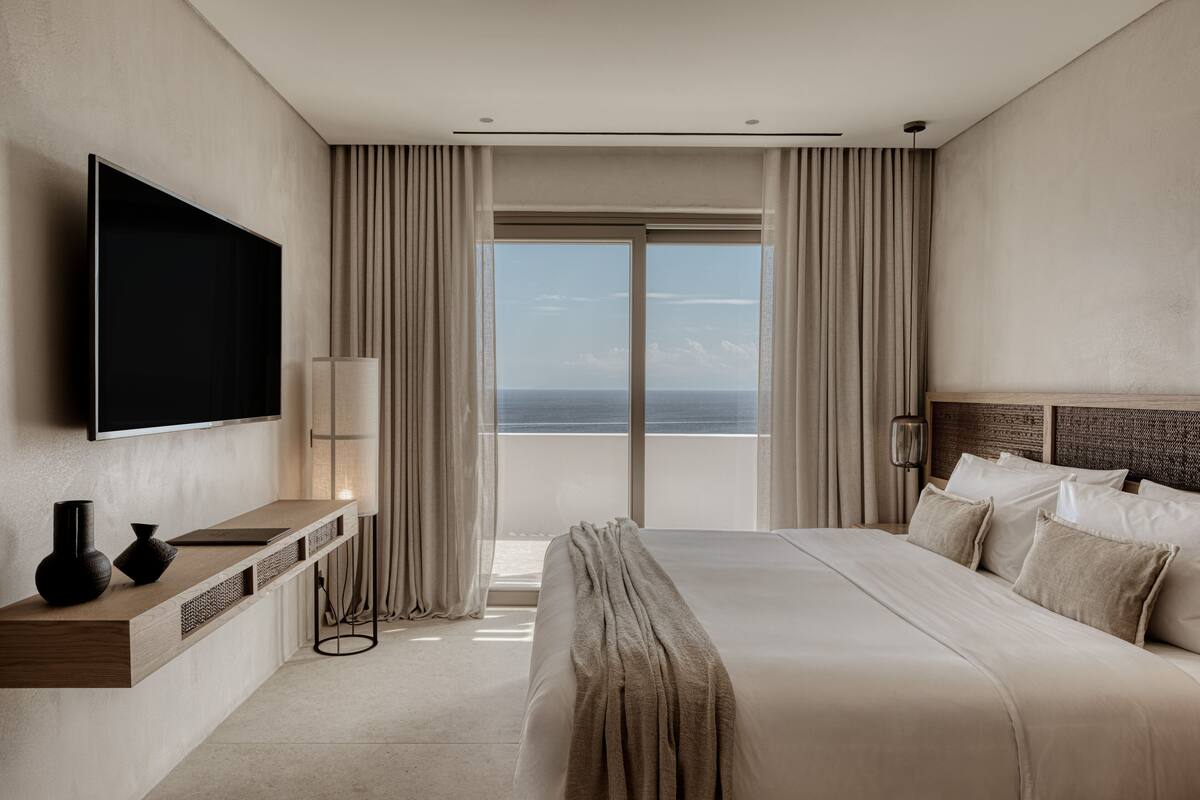 Ein modernes Hotelzimmer mit einem großen Bett, einem Flachbildfernseher, einem Nachttisch mit einer Vase und Meerblick durch raumhohe Fenster.