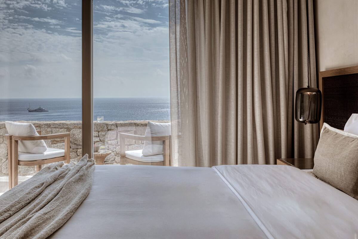 Ein Hotelzimmer mit einem großen Bett, zwei weißen Stühlen und Blick auf das Meer, inklusive Steinwand.