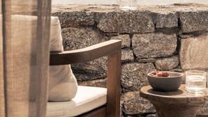 Un mur de pierres, sur une petite table, on aperçoit un saladier rempli de fruits et un verre d’eau. Une chaise en bois avec un coussin est placée à proximité, et de là, on a une vue sur l’océan.