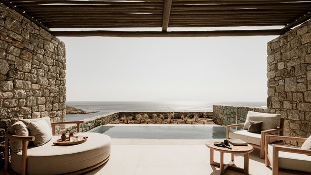 Une piscine à débordement aux murs de pierre, offrant une vue sur l’océan, entourée de mobilier en bois et d’une pergola.