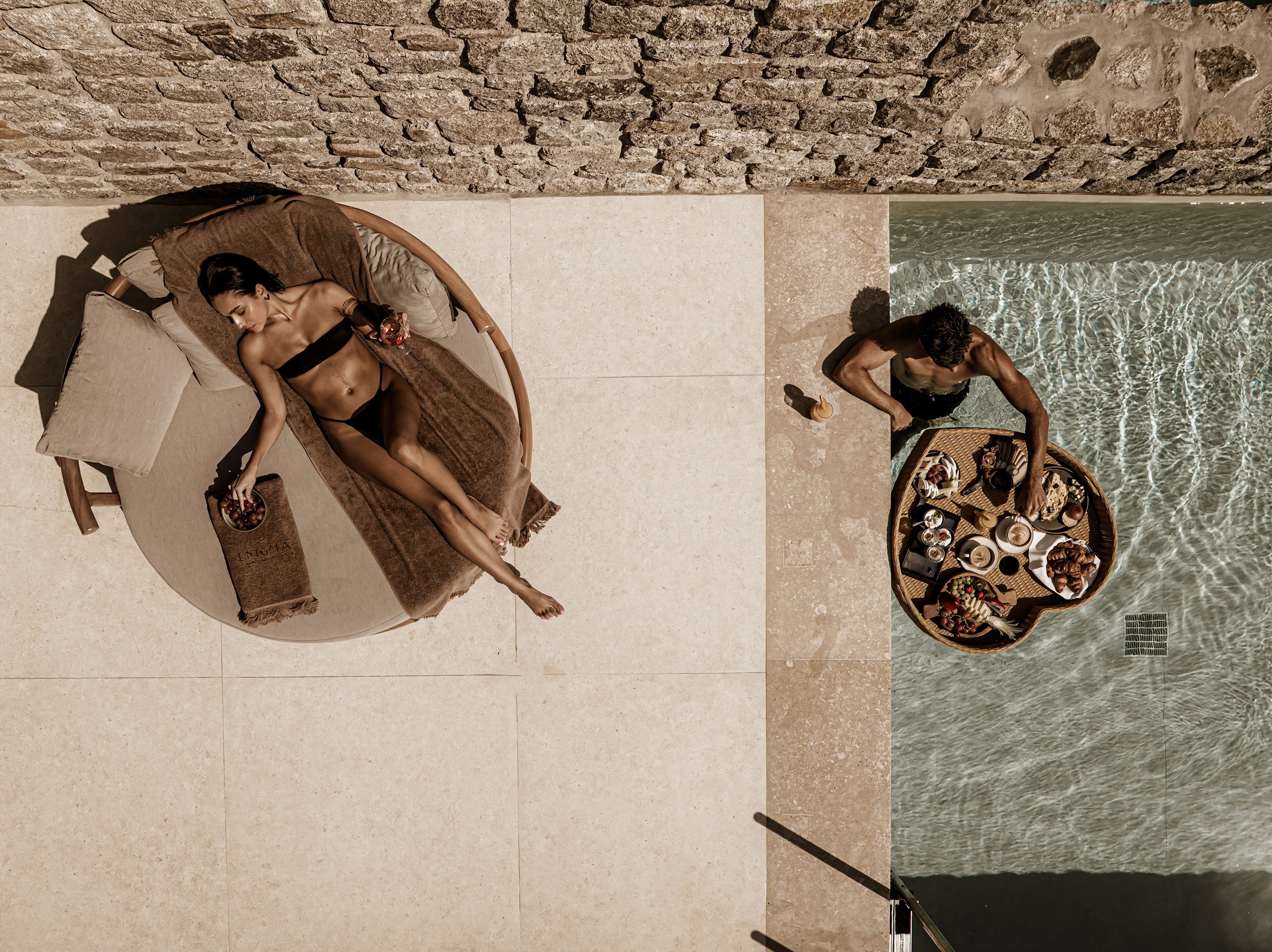 A stone wall and a poolside area with a lounge chair and a dining table with food and drinks.