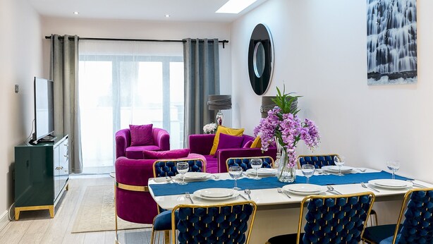 A modern dining area with a table set for four, a large mirror on the wall, and a vibrant purple sofa in the background. - Designer 2-bed 2-bath apartment with a garden (London)