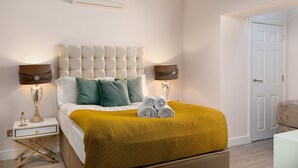 A neatly arranged bedroom with a tufted headboard, two bedside lamps, a bed with a mustard-colored blanket, and a pair of rolled towels. - Designer 2-bed 2-bath apartment with a garden (London)