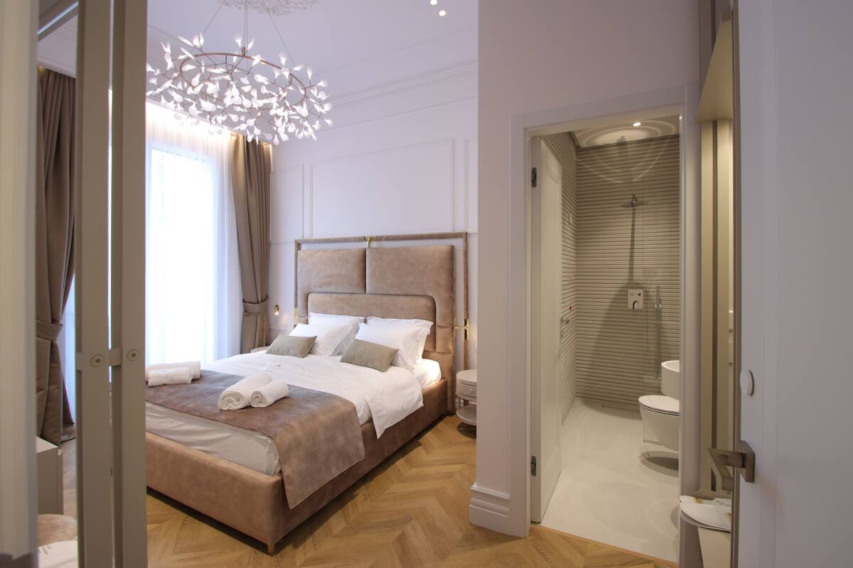Habitación de hotel moderna con una cama grande, un candelabro y un baño visible a través de una puerta abierta.