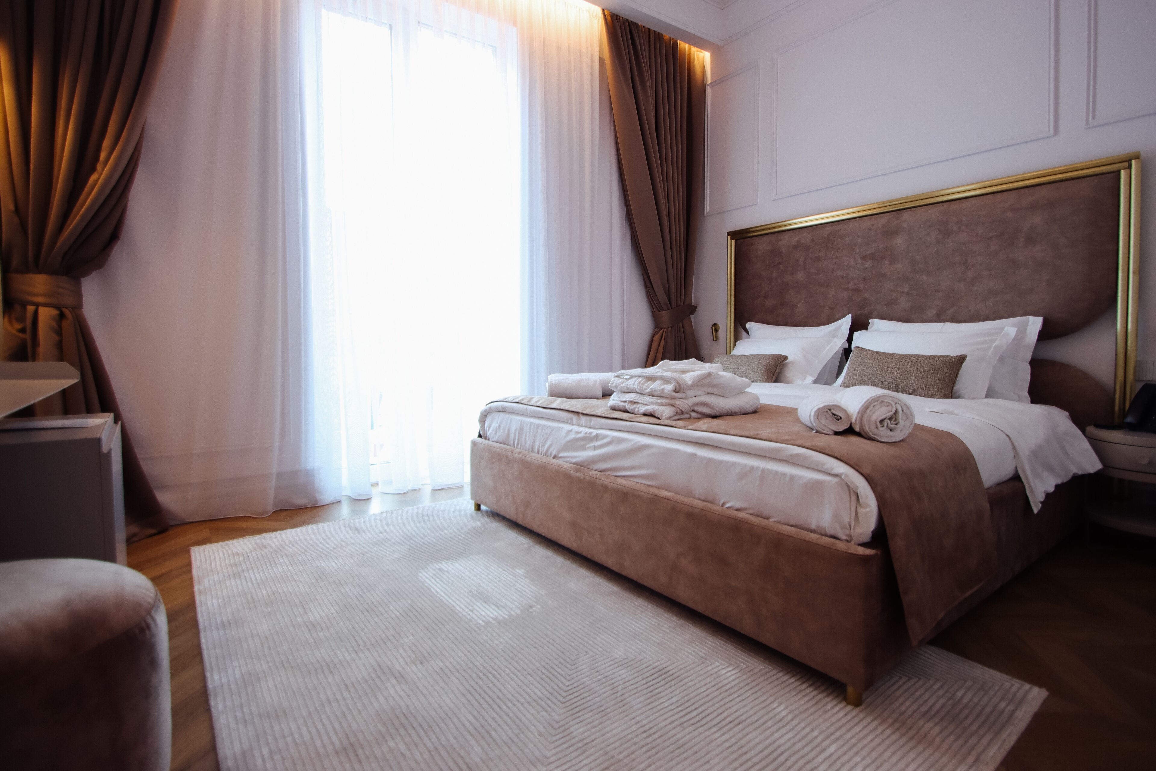 Una cama bien hecha con sábanas blancas y un cabecero marrón, una mesita de noche y un ventanal con cortinas.