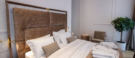 Una cama bien hecha con ropa de cama blanca y cabecera marrón, una mesita de noche con una lámpara, una silla y una planta en maceta en una habitación de hotel.