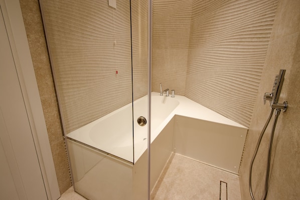 Une salle de bain d’angle avec une douche à parois de verre, une baignoire blanche et un mur beige à la texture particulière.