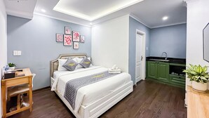 A hotel room with a bed, a desk, a small table, a plant, and a green kitchen area. - Golden Sun Hotel Apartments (Ho Chi Minh City)