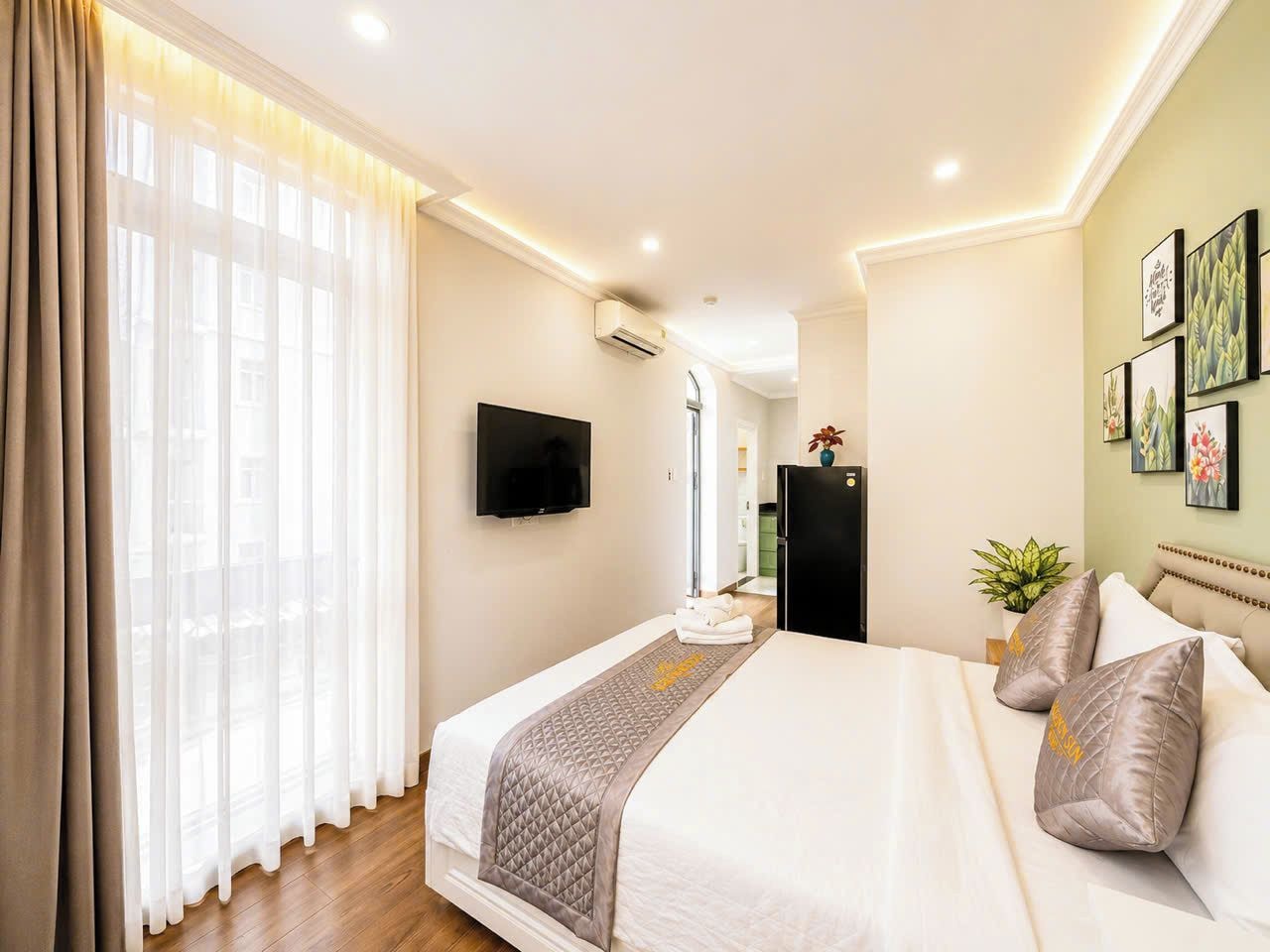 A hotel room with a bed, a TV, a plant, and artwork on the wall.