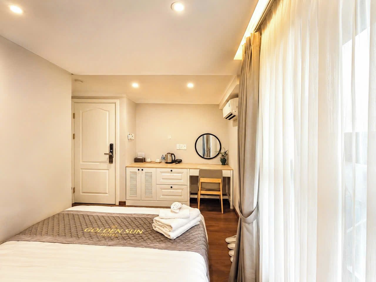 Een hotelkamer met een bed, een bureau en een stoel. Er is een deur, een spiegel en een airconditioning.