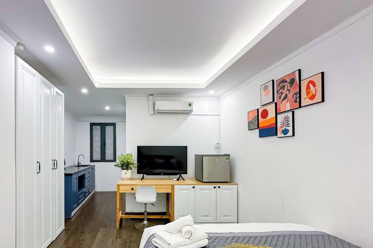 A modern hotel room with a bed, a desk with a TV, a small fridge, a potted plant, and artwork on the wall.