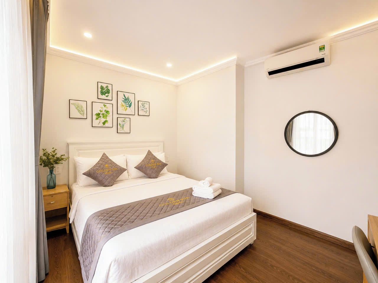 A hotel room with a bed, bedside table, wall-mounted air conditioner, and framed botanical artwork.