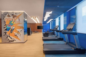 Un gimnàs modern amb equipament d'exercici, un mural gran a la paret i una paret blava amb finestres.