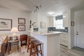 Une cuisine compacte dotée d’un îlot central, de tabourets de bar, d’un coin repas avec une table en osier et d’un grand miroir placé au-dessus de l’îlot.