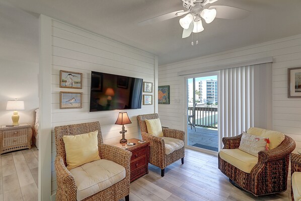 A living room with wicker furniture, a flat-screen TV, and a balcony with a view of buildings.