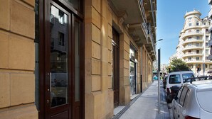 A street view with a building featuring large windows and a door, a sidewalk, parked cars, and a multi-story building in the background. - Gros Arena I | The Rentals Collection (San Sebastián)