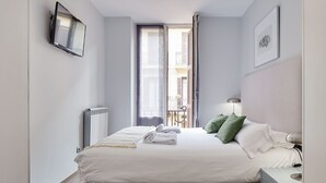 A neatly made bed with white linens and green pillows, a bedside table with a lamp, and a view of a balcony through an open door. - Gros Arena I | The Rentals Collection (San Sebastián)