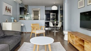 A modern living room with a grey sofa, a round white coffee table, a wooden TV stand, and a dining area with white chairs and a small table. - Gros Arena II | The Rentals Collection (San Sebastián)