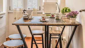 Una cocina con una mesa de madera, dos taburetes, una cafetera, un hervidor, un tarro etiquetado 'CAFÉ' y plantas en macetas.