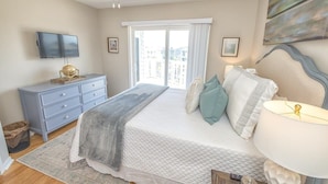 Un dormitorio con cama, cómoda, televisor, ventana con cortinas y un cuadro en la pared.