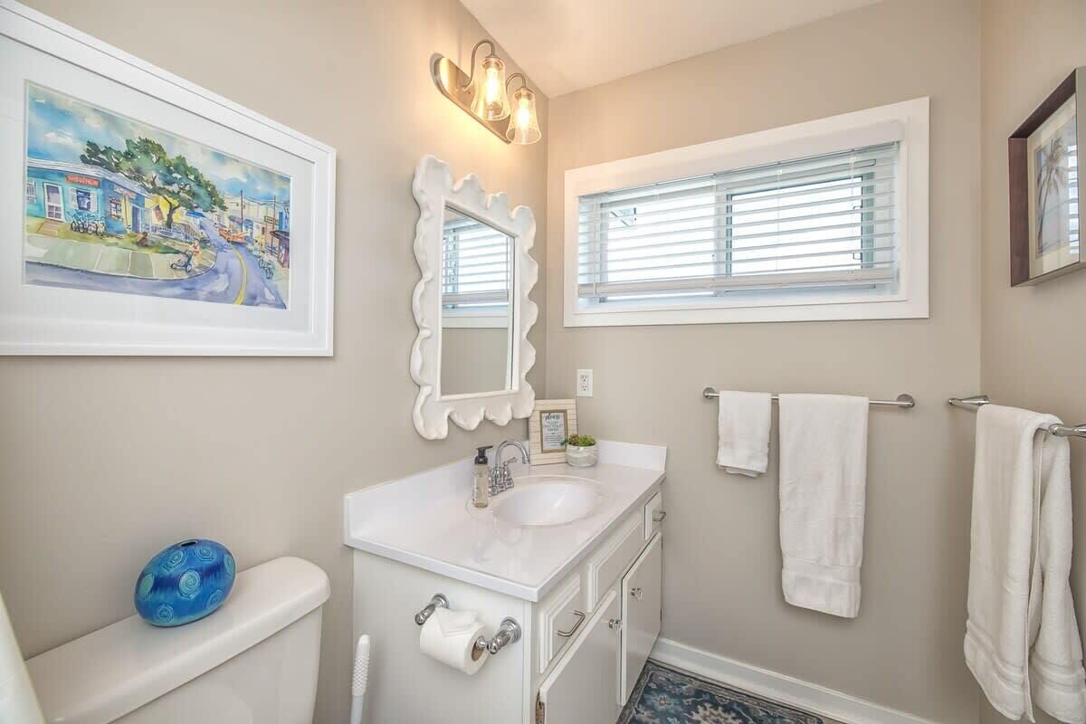 A bathroom with a white vanity, a large mirror, a framed painting, a window with blinds, and a blue decorative item on the toilet.
