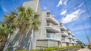 Un edificio frente a la playa con varios balcones, palmeras y un cielo despejado.