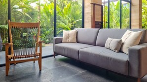 Un salon moderne avec un canapé gris et un fauteuil en bois, de grandes fenêtres offrant une vue sur une végétation luxuriante.