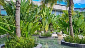 Un bâtiment moderne à la façade vitrée, entouré d’un espace paysager aquatique agrémenté de plantes tropicales.