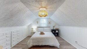 Une chambre avec un plafond en pente, un lit recouvert d’une courtepointe blanche, une table de chevet avec une lampe et une petite étagère ornée d’objets décoratifs.