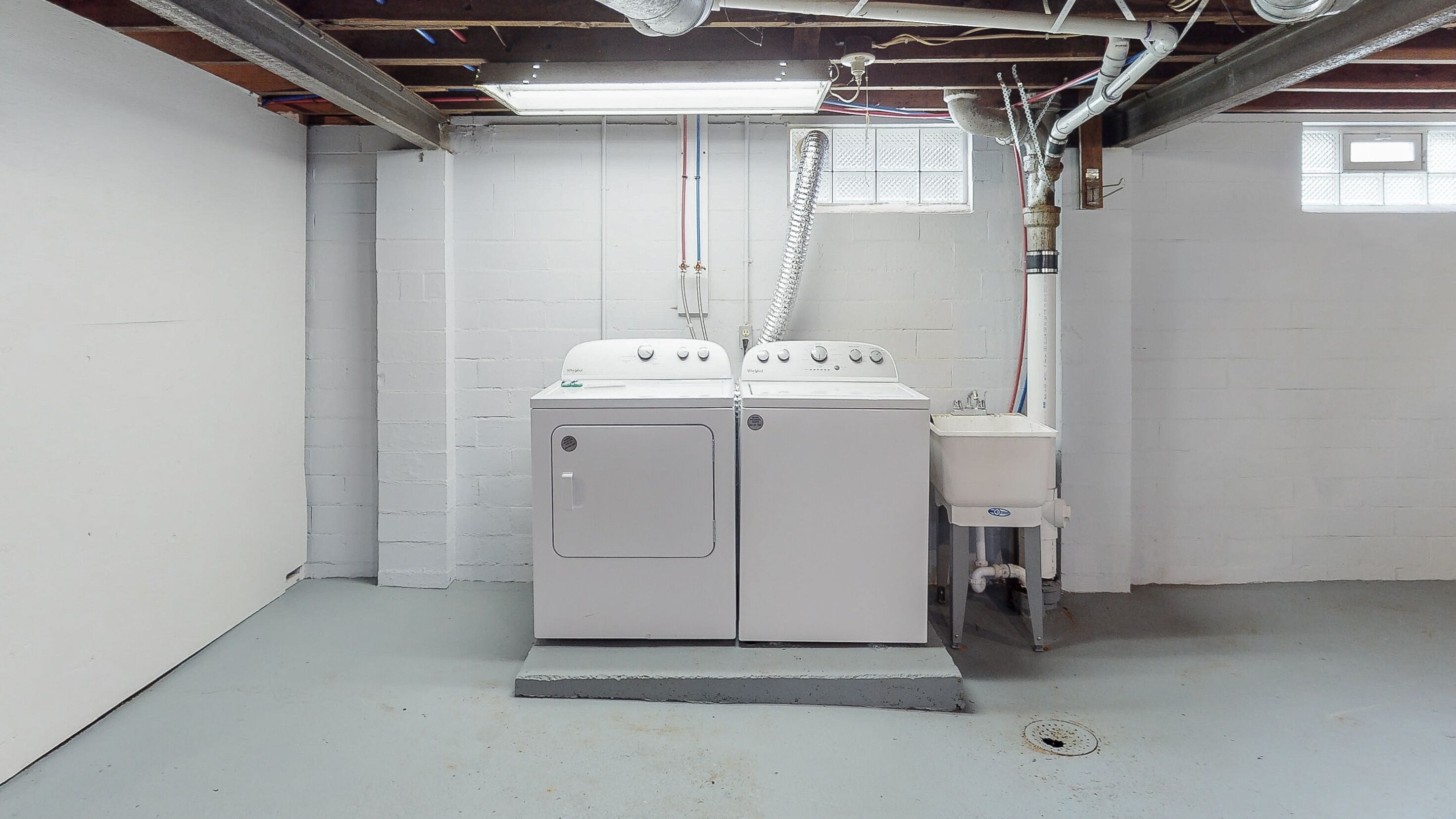 A laundry room with a washing machine and a sink, white walls, and exposed ceiling pipes.