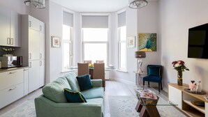 Ein modernes Wohnzimmer mit einem grünen Sofa, einem Esstisch und Stühlen. Ein Fernseher ist an der Wand montiert und eine Pendelleuchte hängt von der Decke.