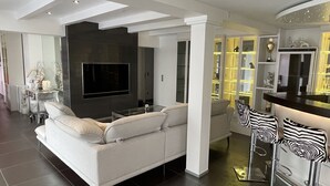 Una sala de estar moderna con un televisor de pantalla plana grande, un sofá blanco y una zona de bar con sillas a rayas de cebra.