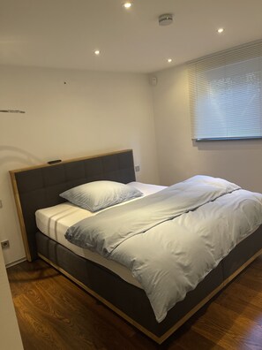 Un dormitorio moderno con cama, suelo de madera y ventana con persianas.