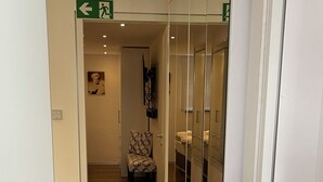 Un pasillo con un muro espejado, una silla y una cama a lo lejos.