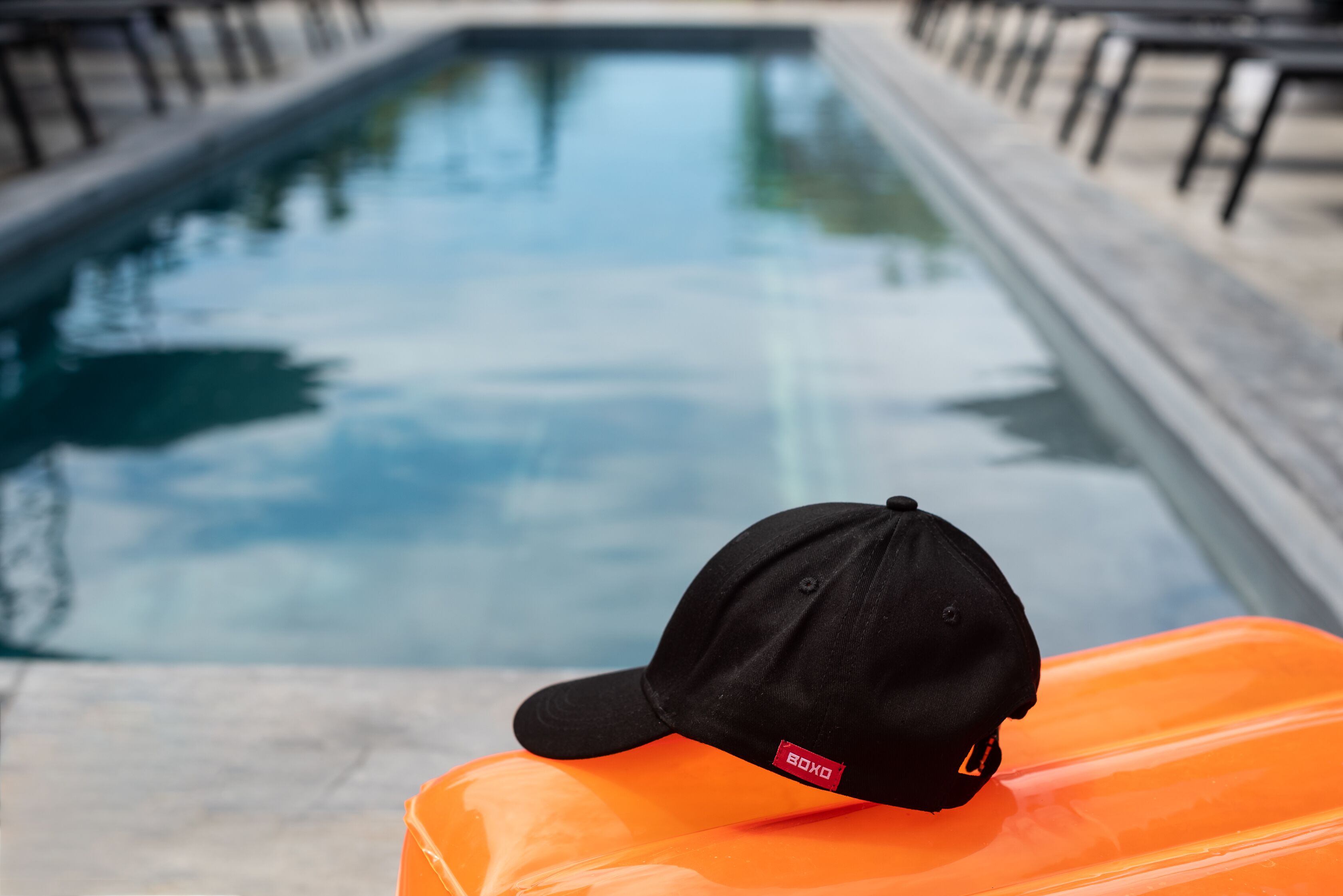 Una gorra negra sobre un objecte taronja a la vora d'una piscina, amb un cel blau clar al fons.