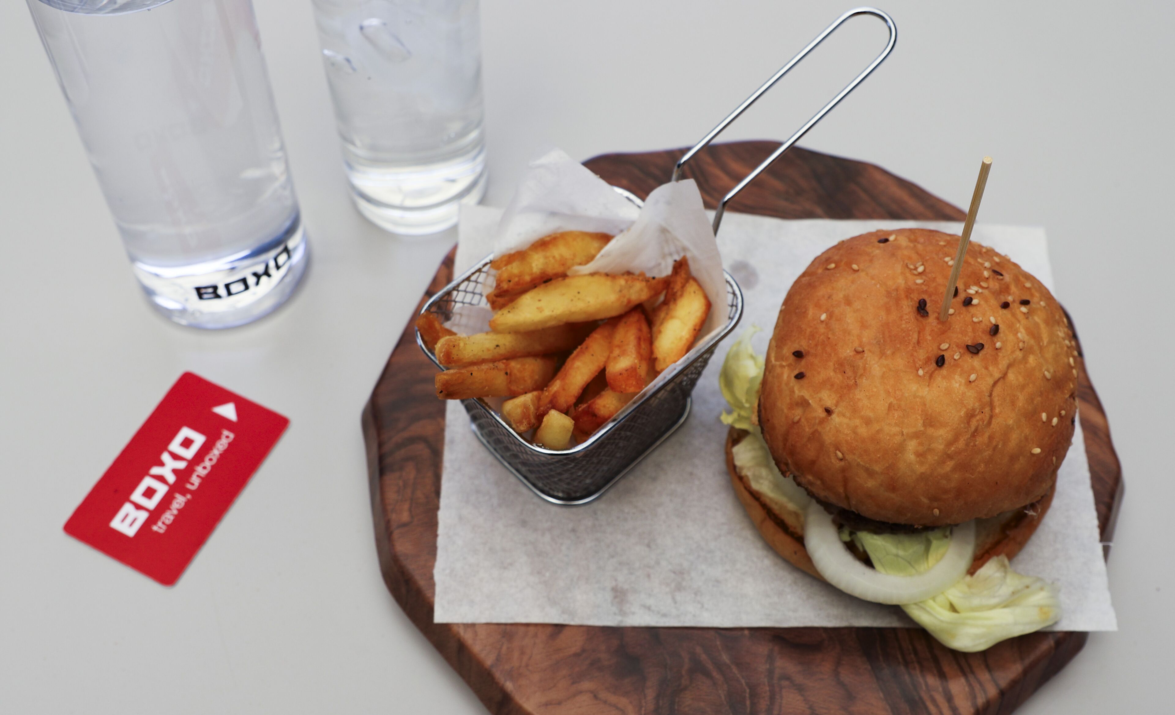 Un hamburger et des frites servis sur une planche en bois, accompagnés d’une boisson et d’une carte placés à côté.