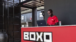 A man in a red shirt standing behind a counter with a laptop, with a sign that reads "BOXO" on the wall. - Boxo Diani (Diani Beach)