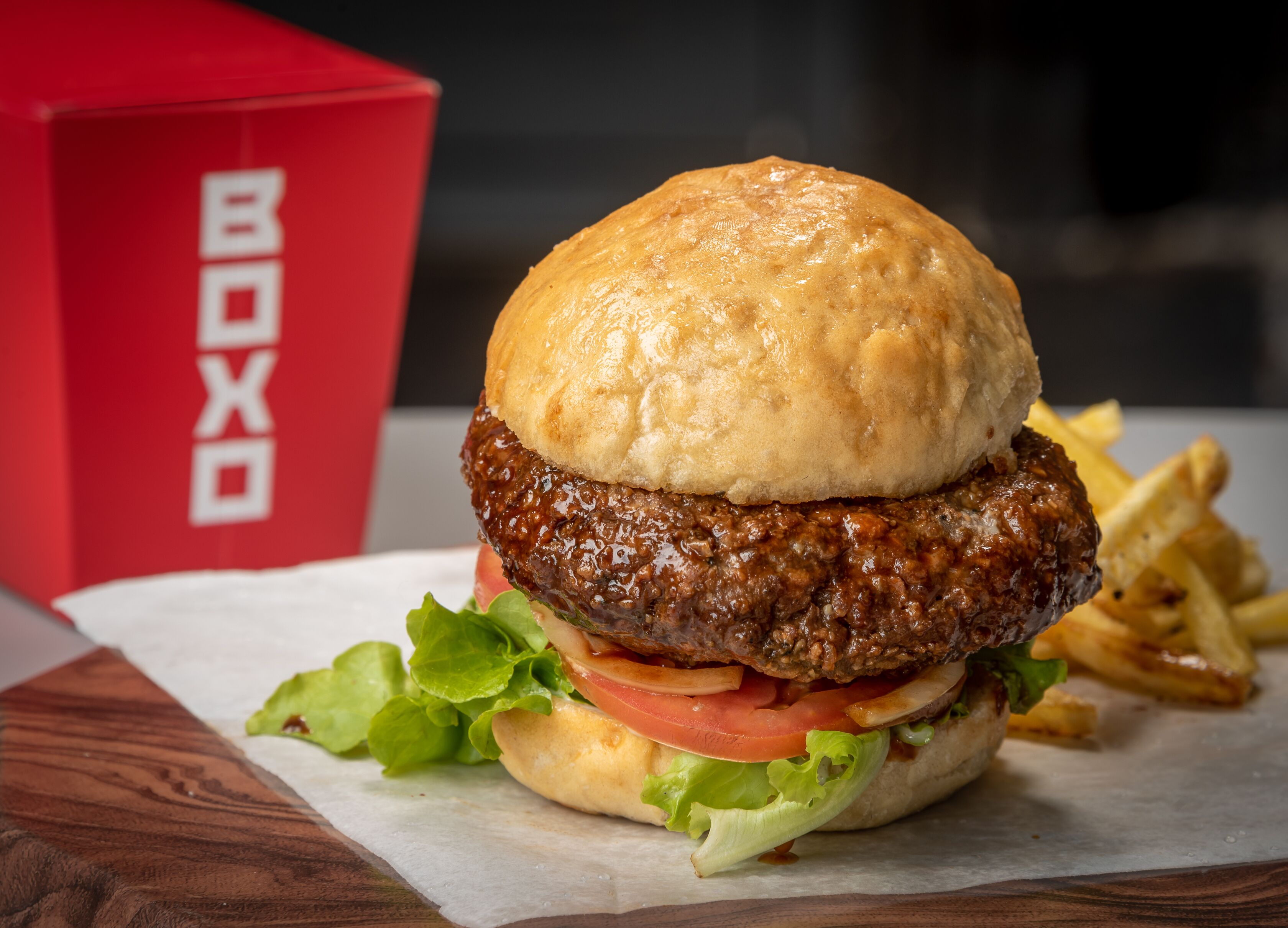 A burger with lettuce, tomato, and a bun, accompanied by fries, on a wooden table with a red box labeled "BOXO" in the background.