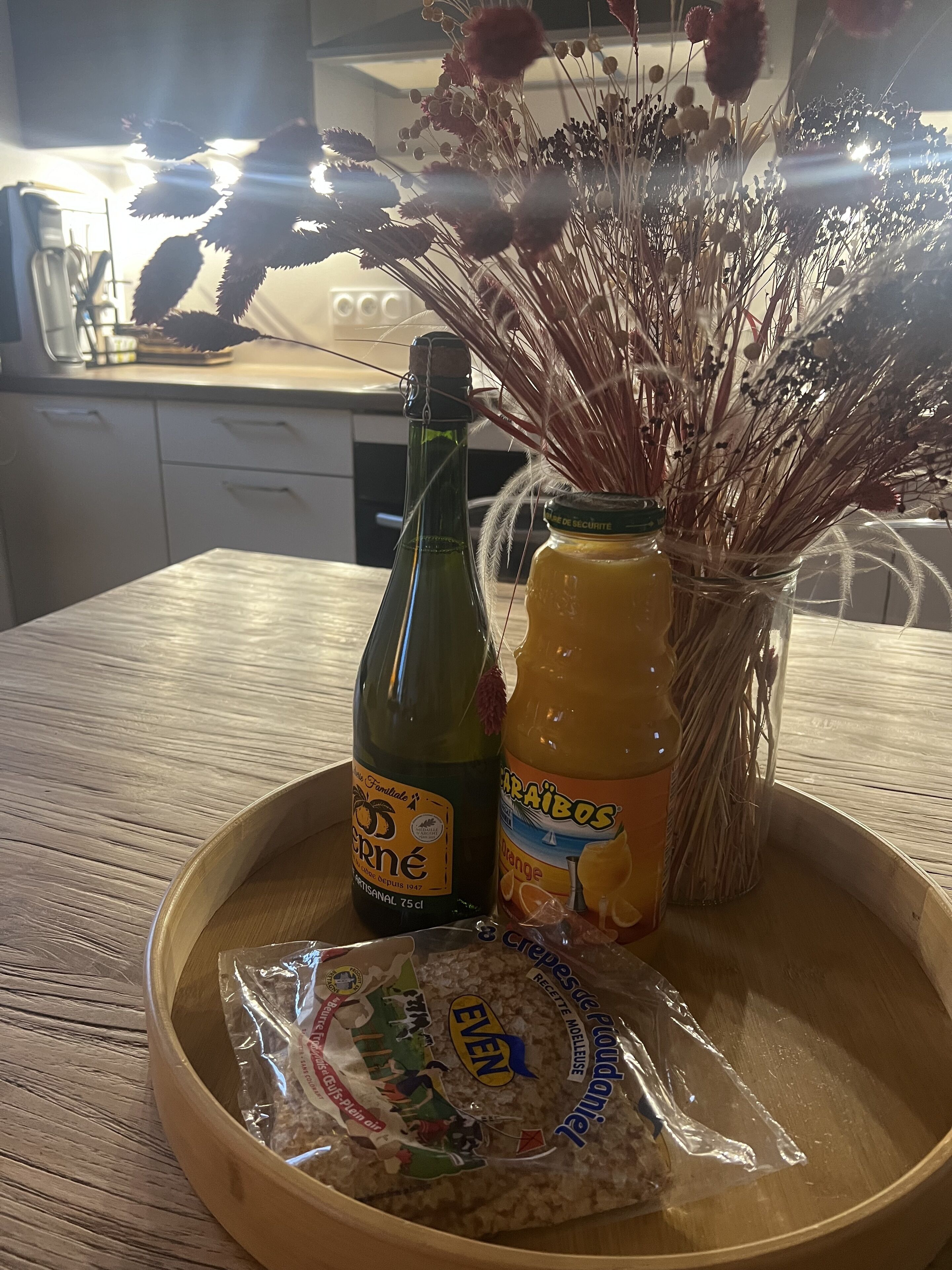Ein Holztablett mit einer Flasche Perné, einer Flasche Orangensaft und einem Laib Brot der Marke Even auf einer Küchenarbeitsplatte.