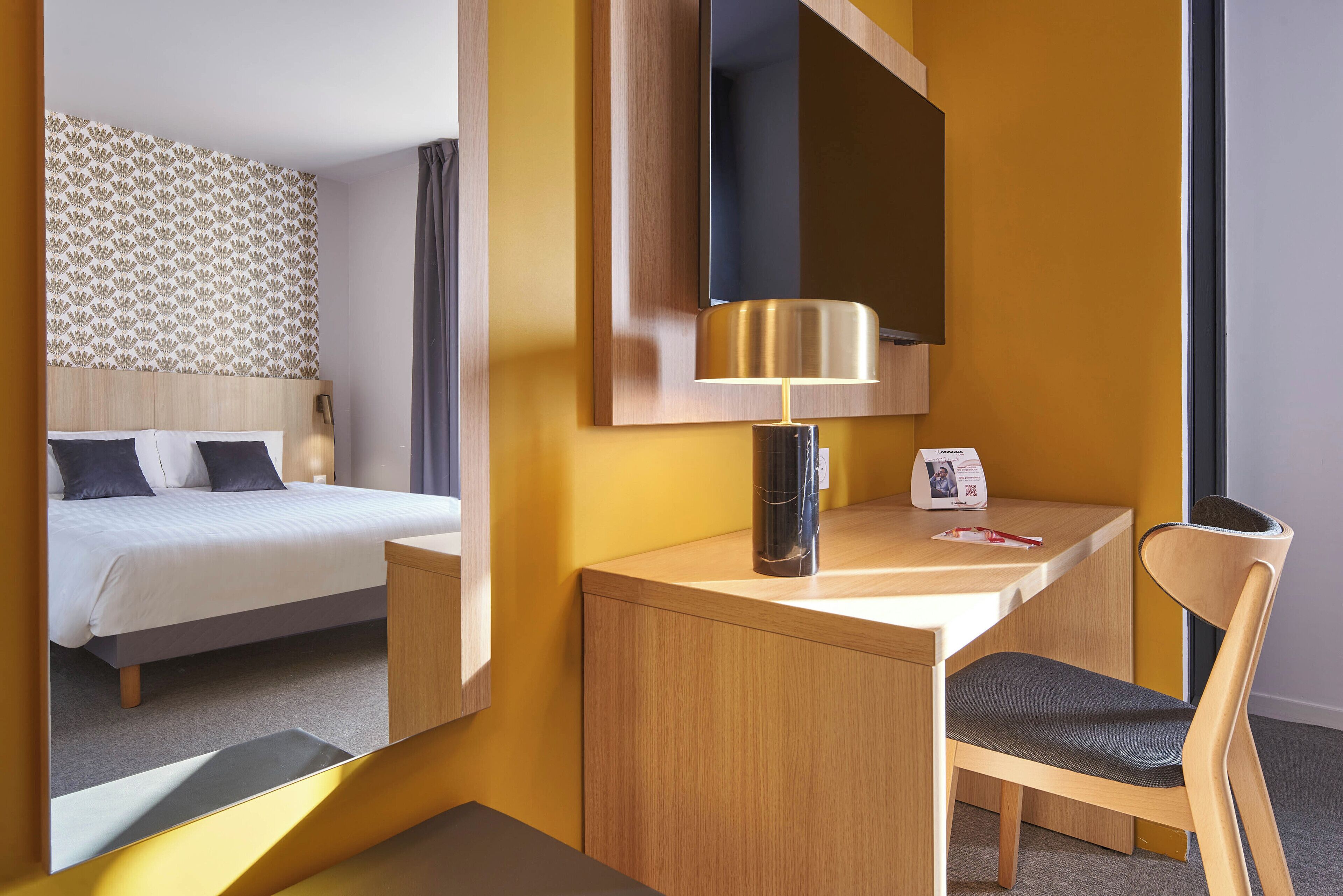 Et hotelværelse med en seng, et skrivebord med lampe, en stol og et stort spejl.