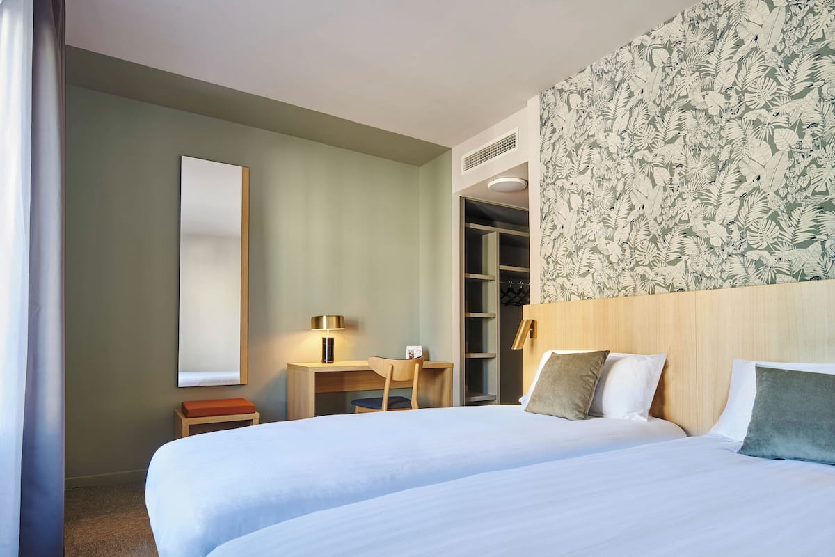 A hotel room with two beds, a desk, a chair, a wardrobe, and a large mirror.