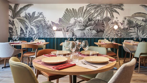 A restaurant with round tables, chairs, and a mural of tropical foliage on the wall. - Logis Hôtel Larivaut, Puy-Guillaume (Puy-Guillaume)