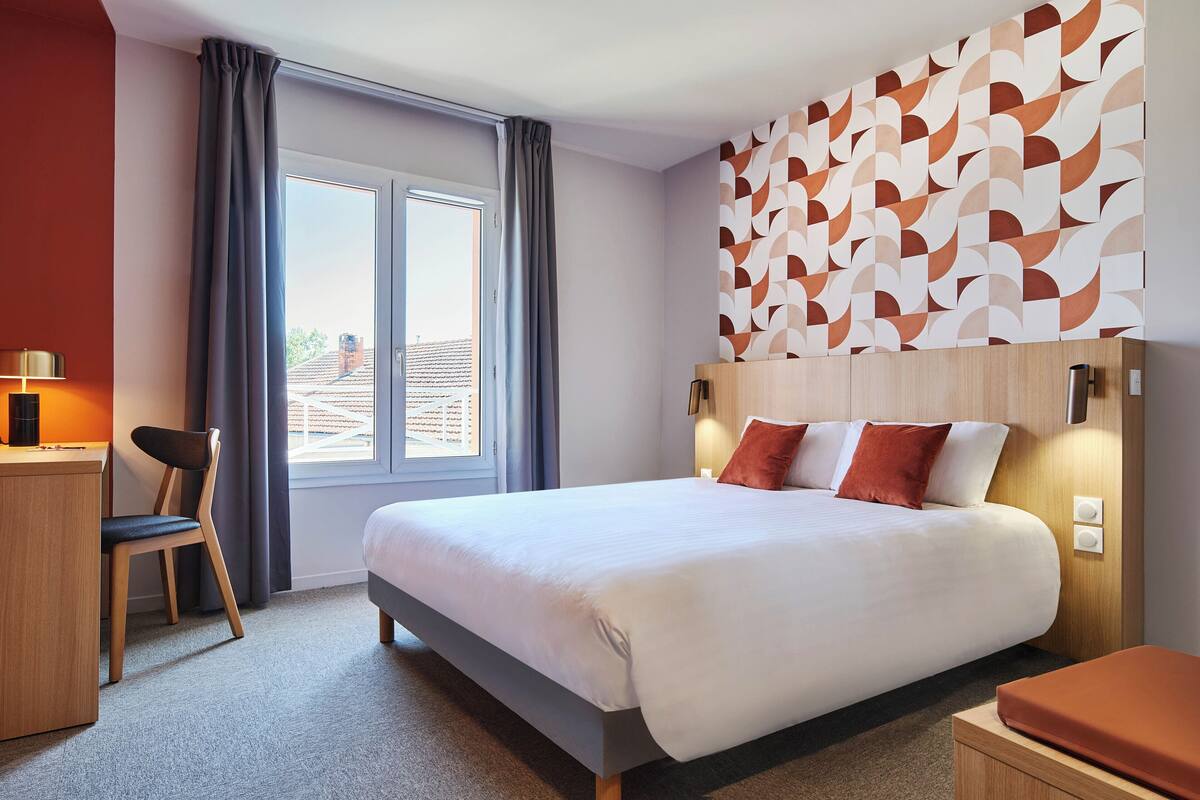 A modern hotel room with a large bed, a desk, and a chair. There is a window with curtains, a patterned wall, and a view of buildings outside.