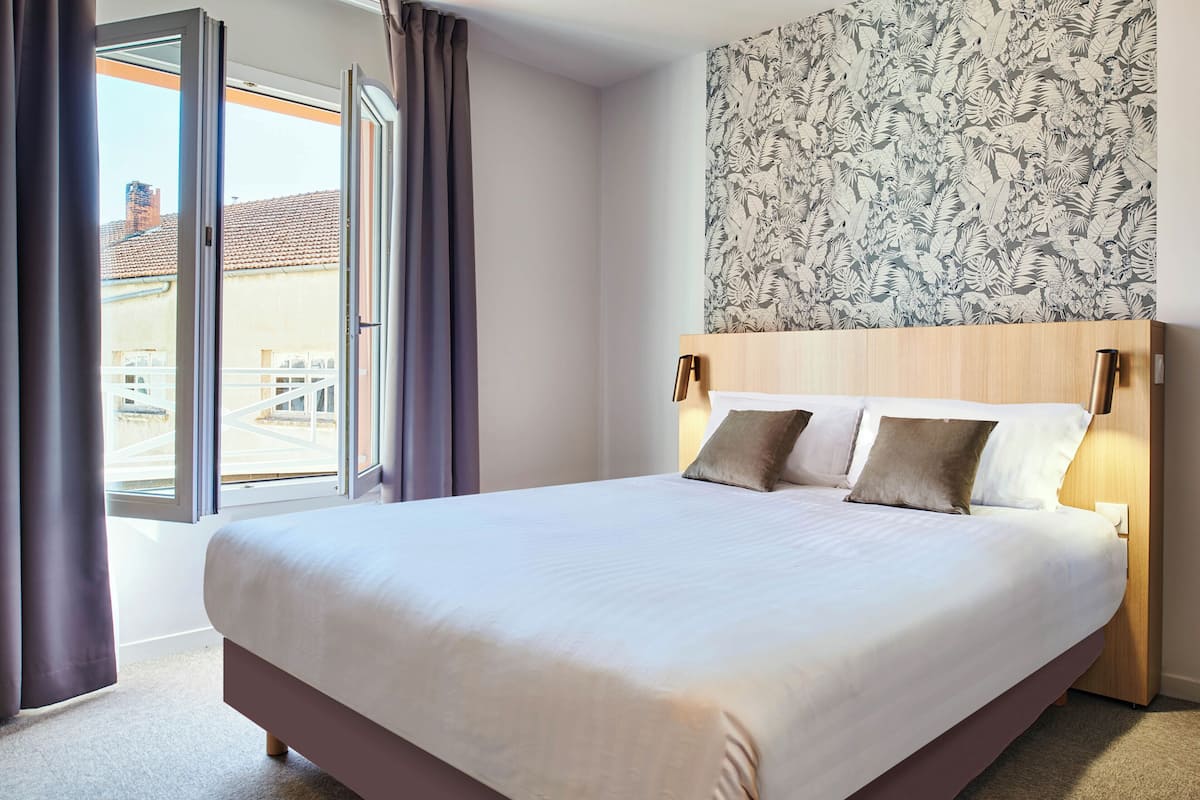 A hotel room with a large bed, a wooden headboard, and a view through a sliding door to the outside.